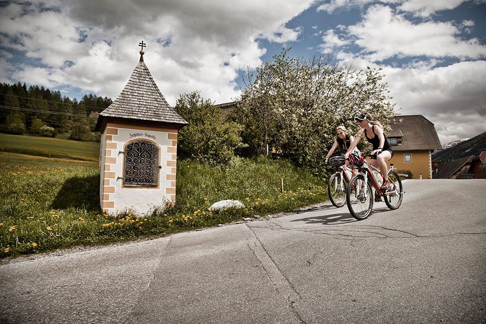 Two cyclists pass a wayside shrine near a 3-star family pension in Mauterndorf, surrounded by lush mountains.