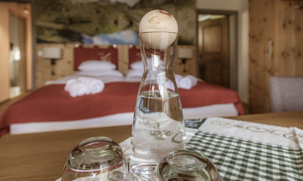 Gemütliches Zimmer mit Holzdekor, roter Bettdecke, Wasserkaraffe, Bergwandbild in einem Alpengästehaus.