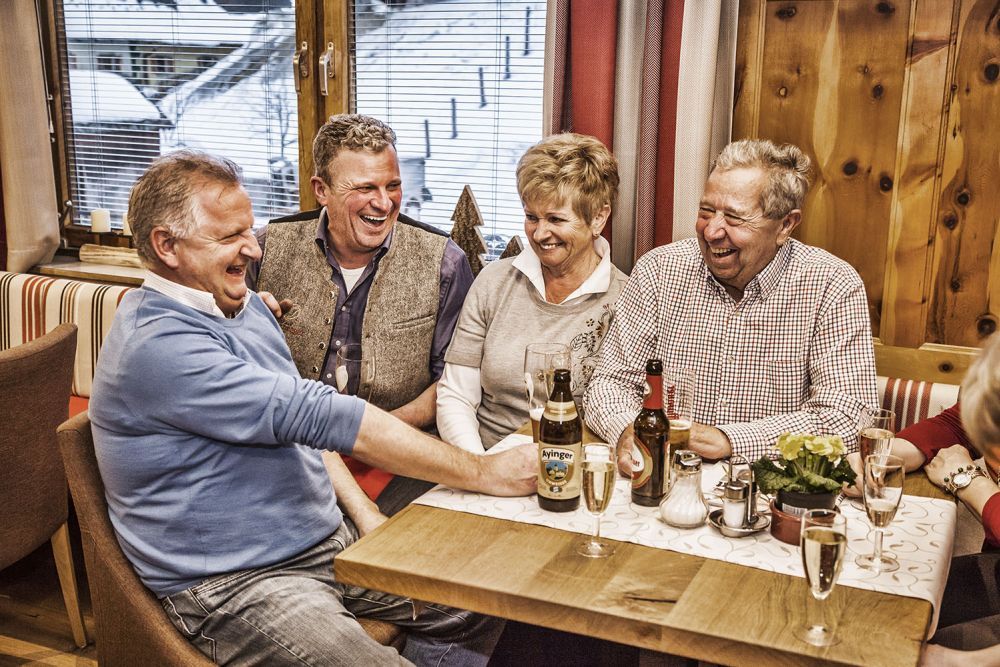 Vier Erwachsene lachen bei Getränken in einer gemütlichen Alpenpension.