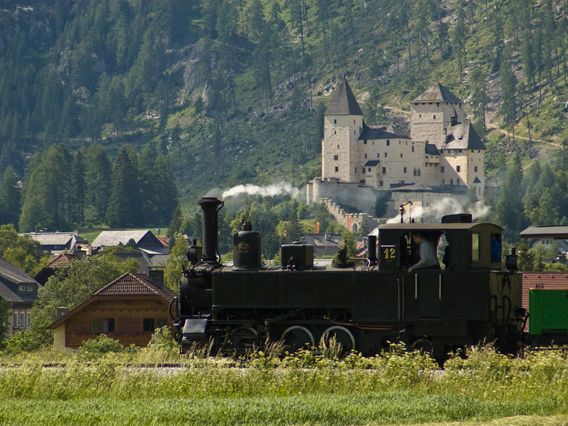 Historische Lokomotive fährt vor einer Burg inmitten grüner Berge.