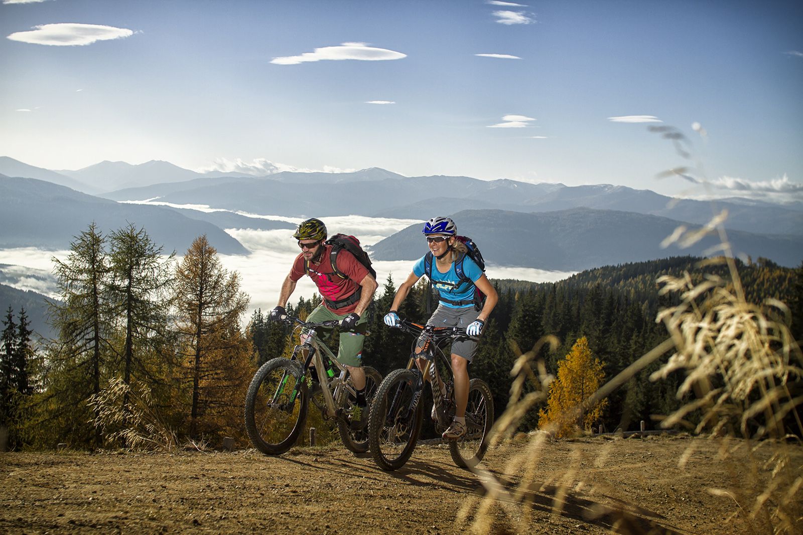 Zwei Radfahrer auf Bergpfad mit nebligem Tal und Gipfeln im Hintergrund.