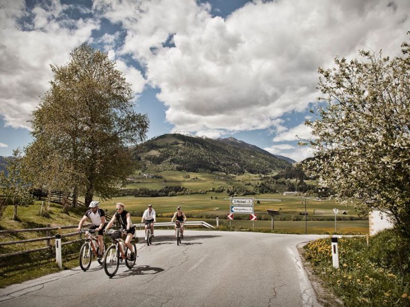 Radfahrer auf ländlicher Straße im Salzburger Lungau, Berge und grüne Landschaft unter wolkigem Himmel.
