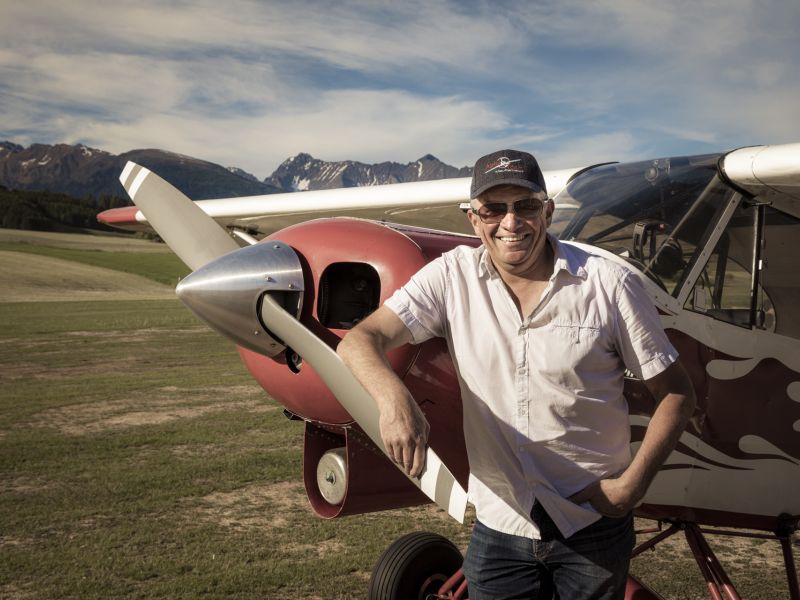 Lächelnder Mann lehnt an einem Oldtimer-Flugzeug, Berge im Hintergrund.